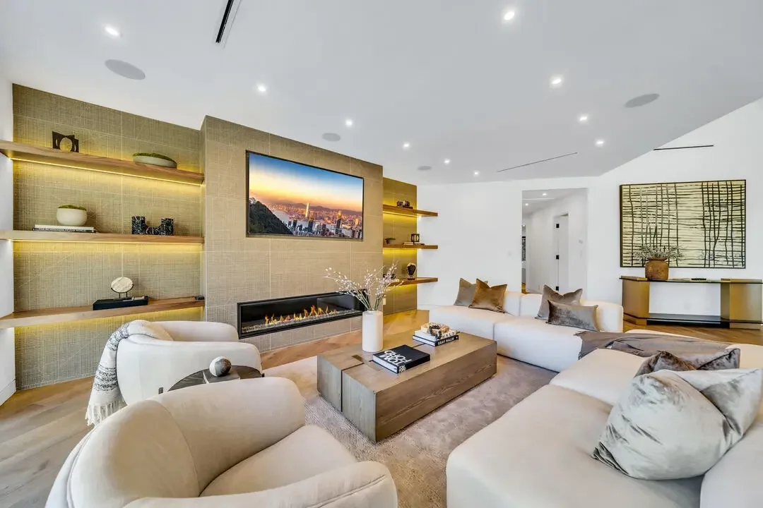 Modern living room with a large L-shaped white sofa, beige armchairs, a wooden coffee table with books and a vase, a wall-mounted TV, decorative shelving with vases and decor, a linear fireplace, and contemporary artwork on the walls.