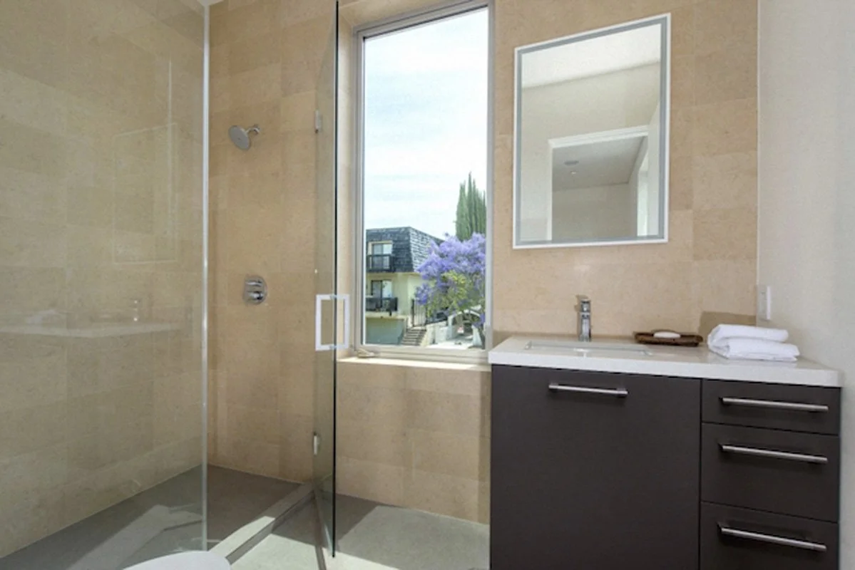 Contemporary bathroom in West Hollywood residence with glass shower, stone tile walls, and large window.