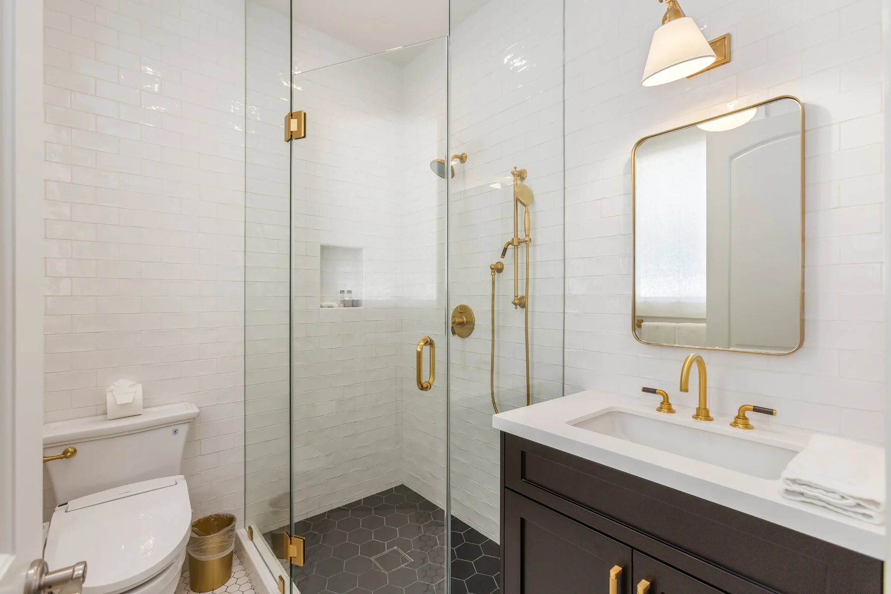 Bathroom in a custom Brentwood home featuring a glass-enclosed shower with white subway tile, brass fixtures, black vanity with white countertop, and hex tile flooring.