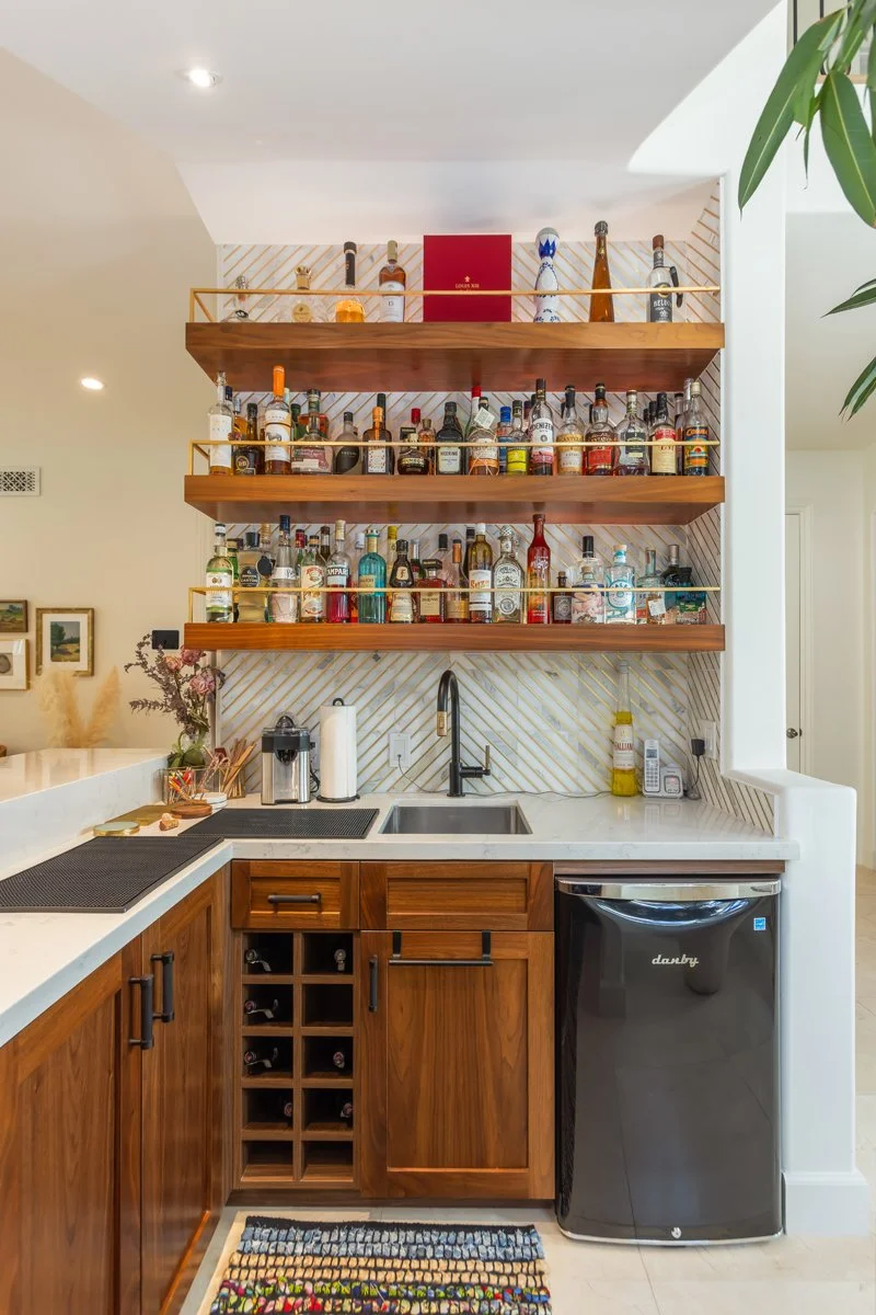 Built-in bar area in a custom Brentwood home featuring walnut cabinetry, open wood shelving with liquor display, marble countertop, integrated sink, wine storage, and compact beverage fridge.