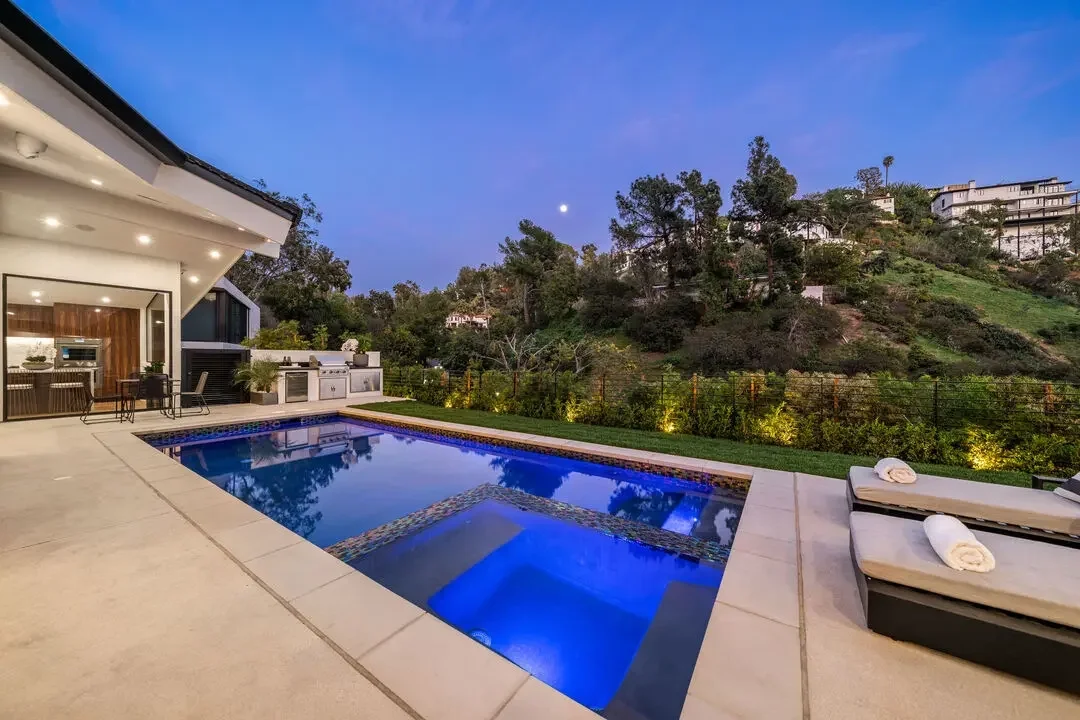 Modern backyard with a swimming pool, outdoor kitchen, and lounge chairs, overlooking a hillside with trees and houses, under a twilight sky.
