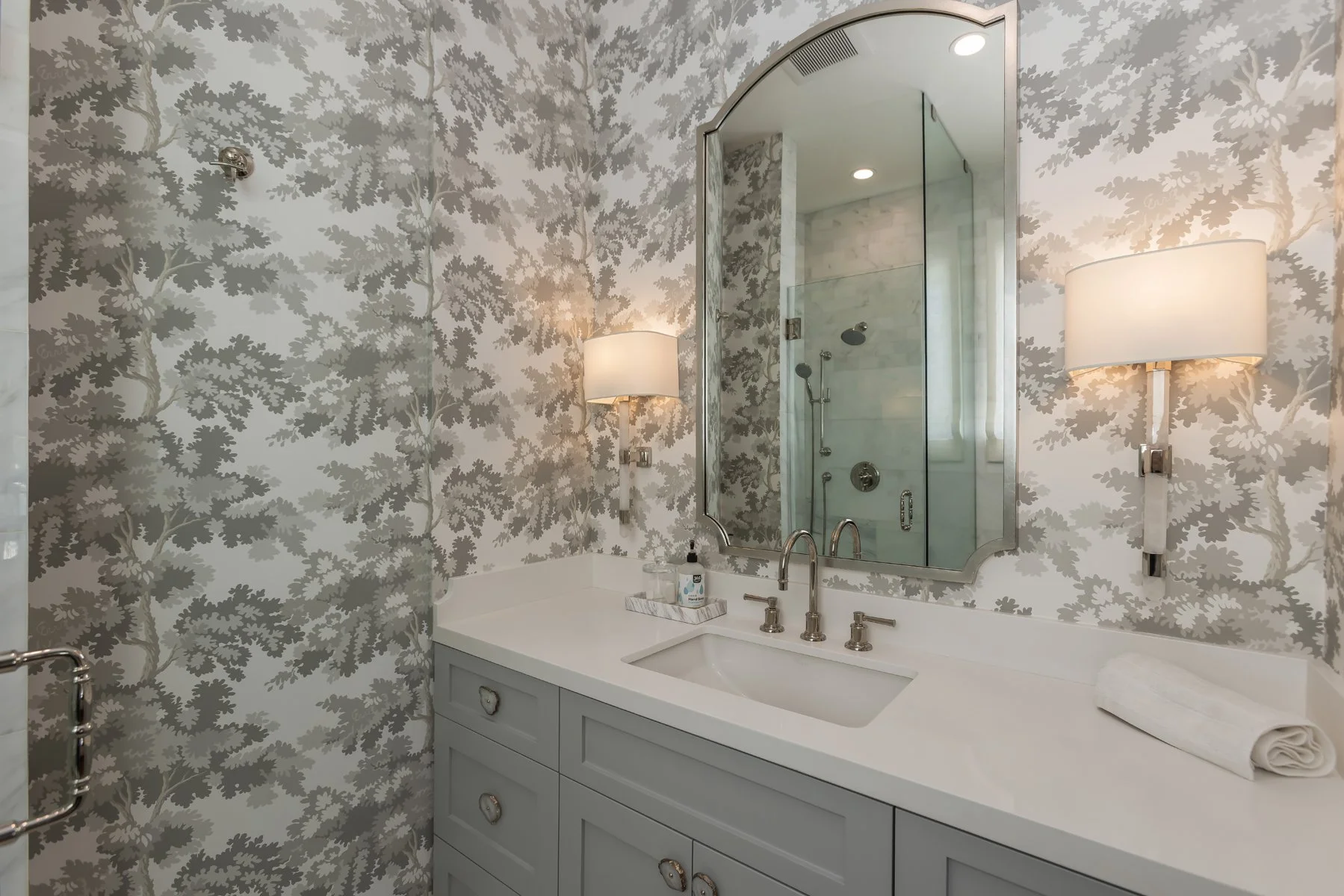 Bathroom vanity in a custom Brentwood home featuring soft gray cabinetry, white countertop, patterned wallpaper, wall sconces, and an arched mirror.