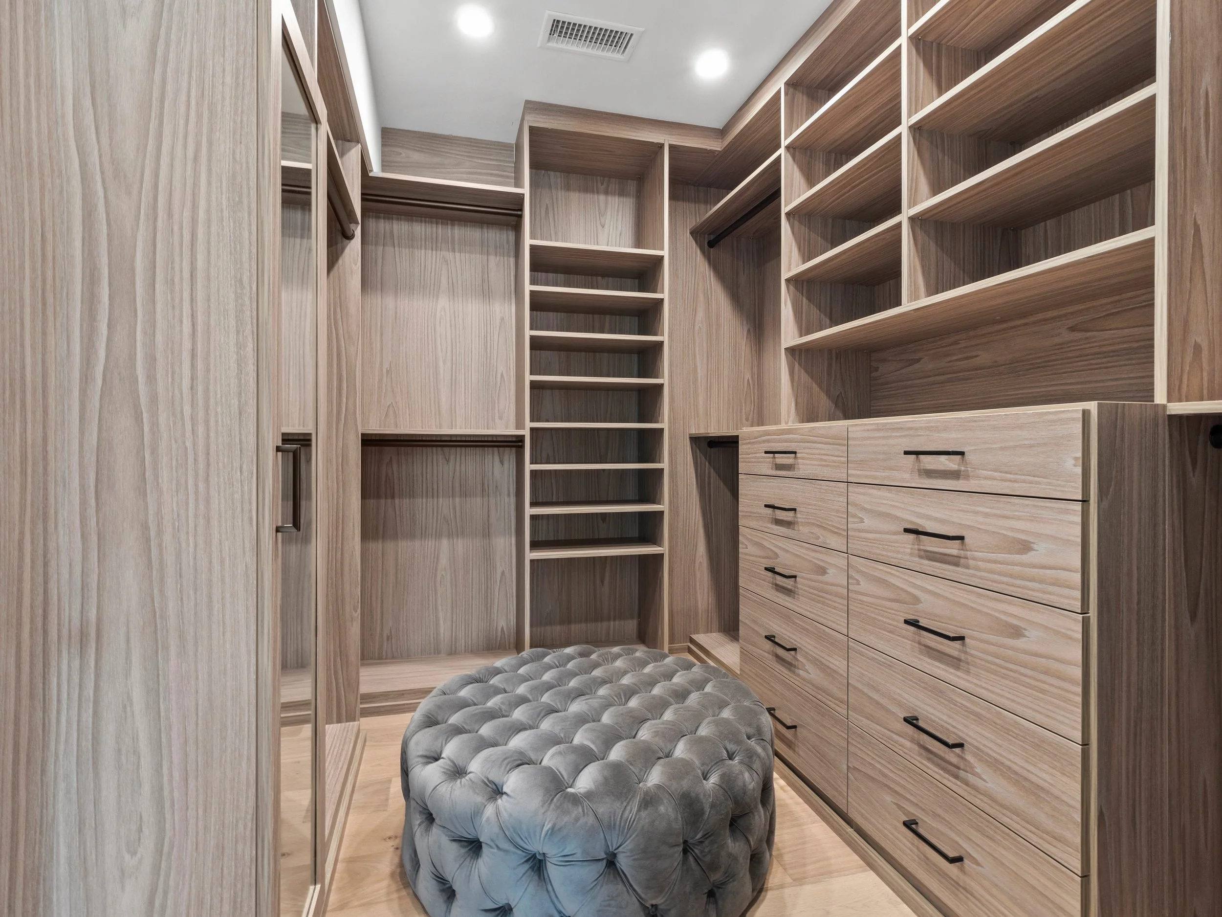 Hancock Park custom walk-in closet with warm wood built-ins, open shelving, drawer storage, hanging space, and tufted upholstered ottoman.
