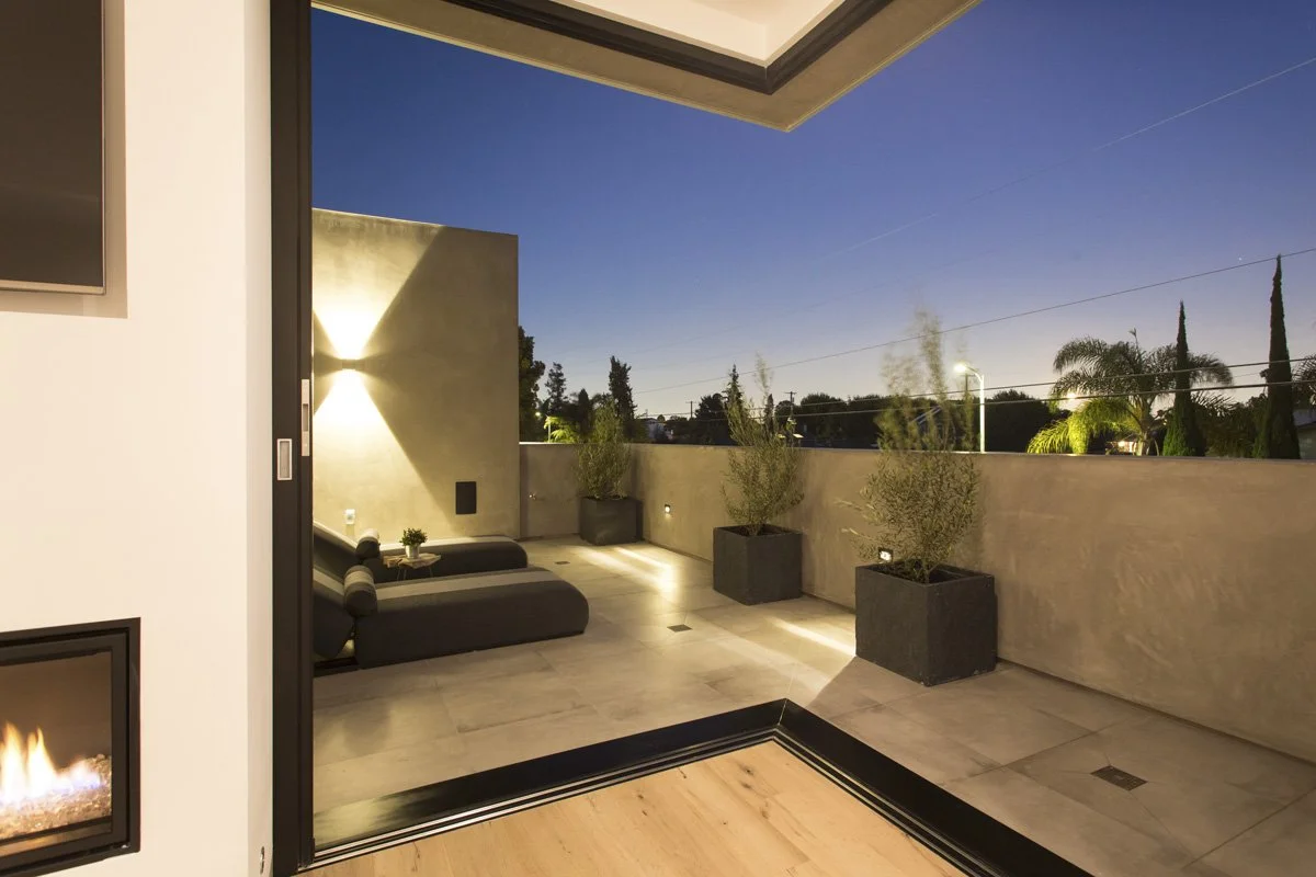 Indoor-outdoor primary suite with sliding glass door, private terrace seating, textured stone pavers, and sculptural wall lighting detail.