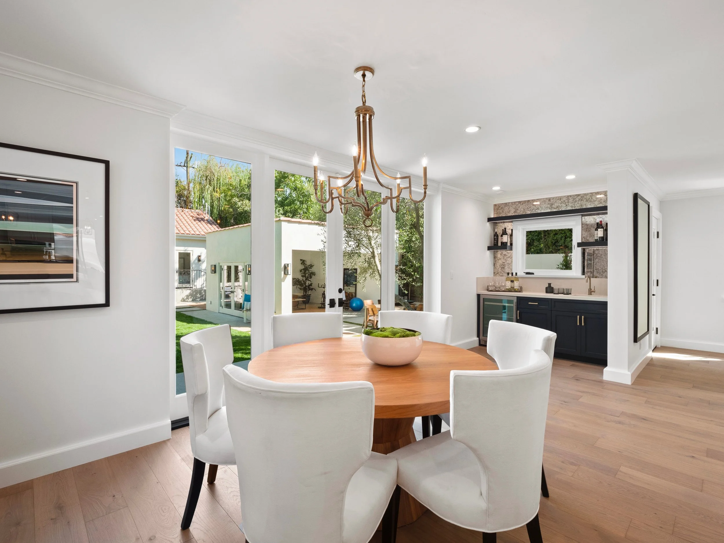 Hancock Park dining room with round wood table, white upholstered chairs, brass chandelier, and glass doors opening to a landscaped courtyard with guest house beyond.