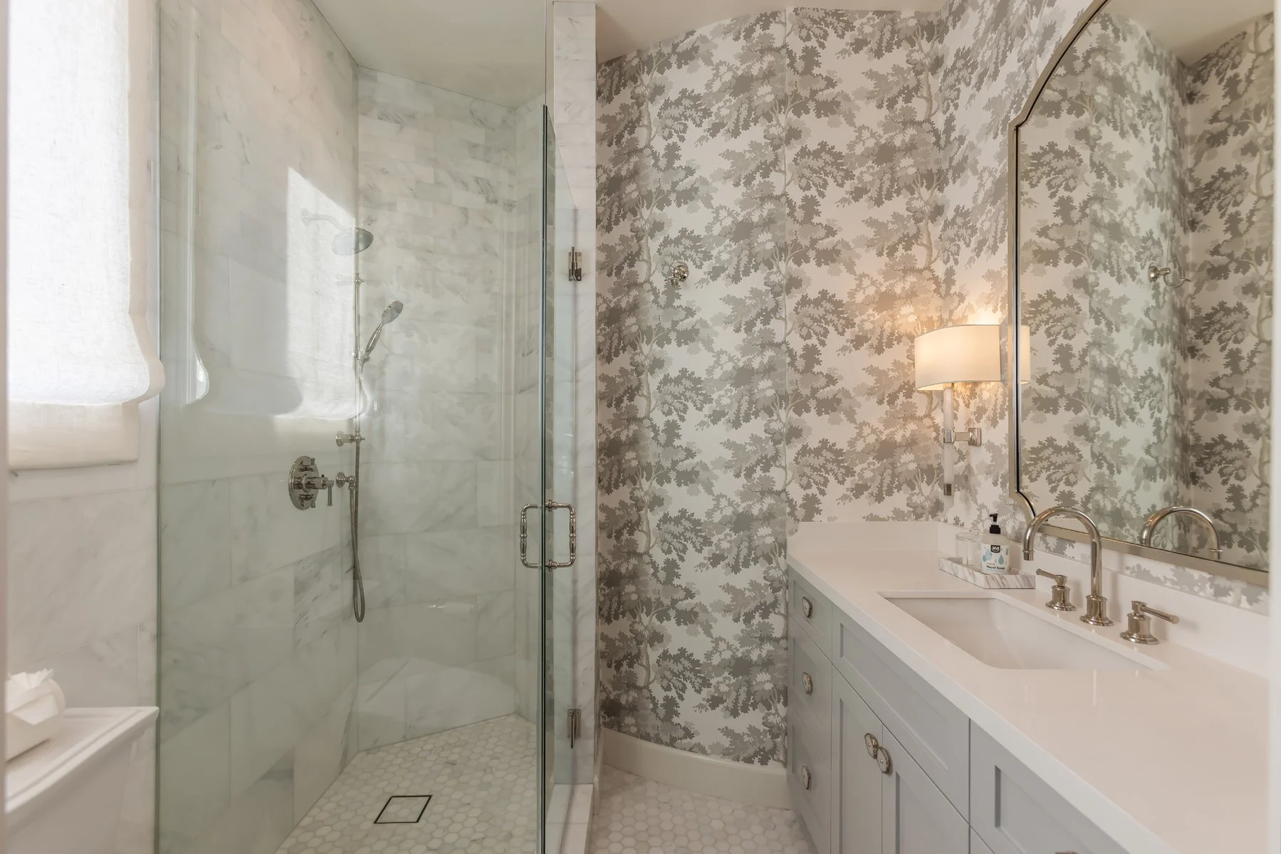 Primary bathroom in a custom Brentwood home featuring a marble-tiled shower, glass enclosure, soft patterned wallpaper, light blue vanity, and classic chrome fixtures.