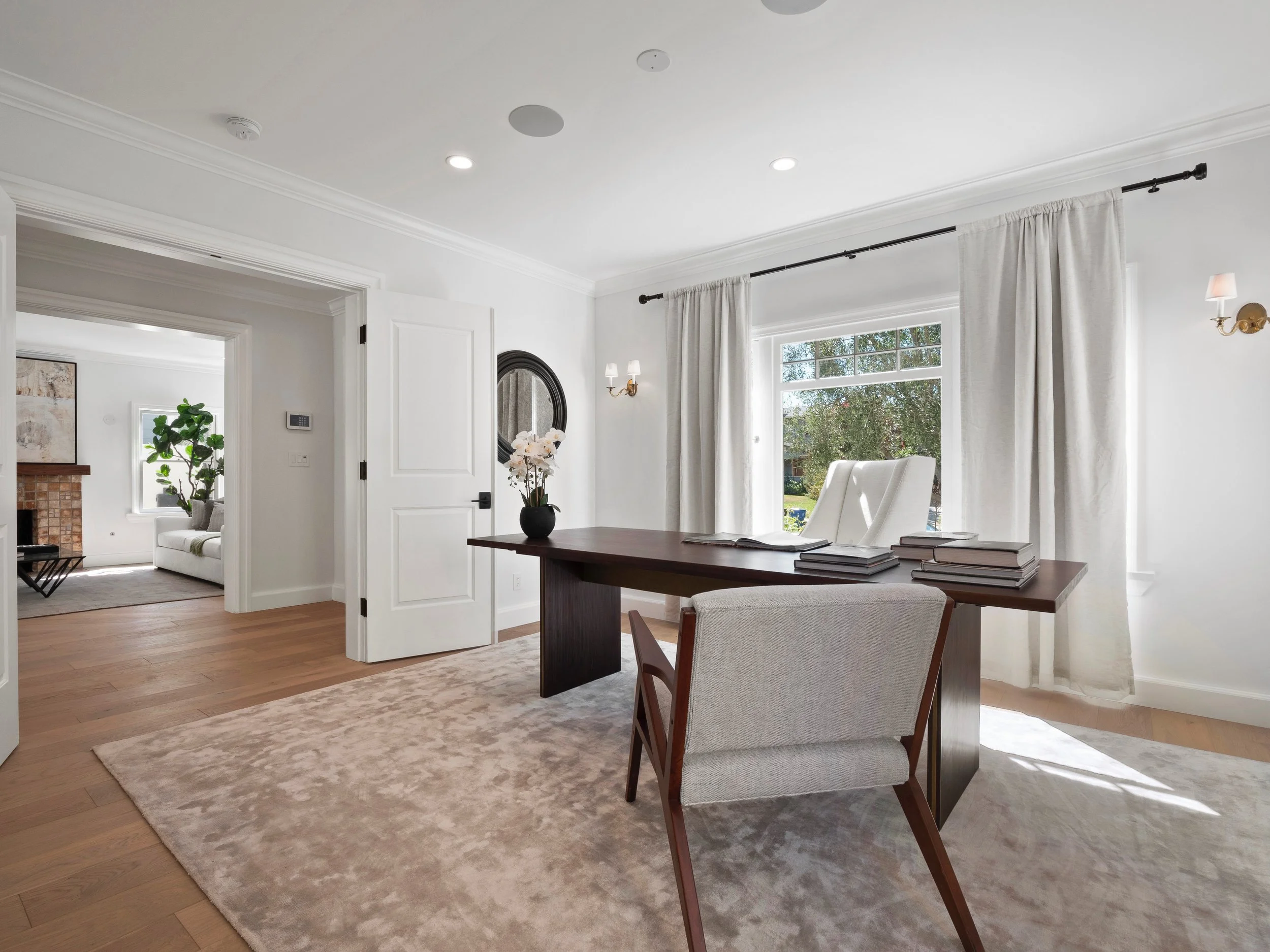 Hancock Park home office with dark wood desk, upholstered chairs, soft neutral rug, white walls, brass sconces, and large window overlooking the garden.