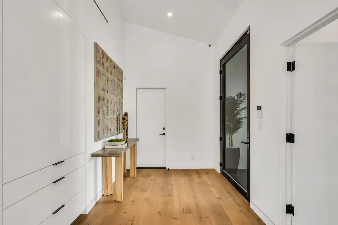 Minimalist interior hallway with white walls, wooden floors, a small wooden table with decor, abstract wall art, a glass door with a plant behind it, and a closed door at the end.