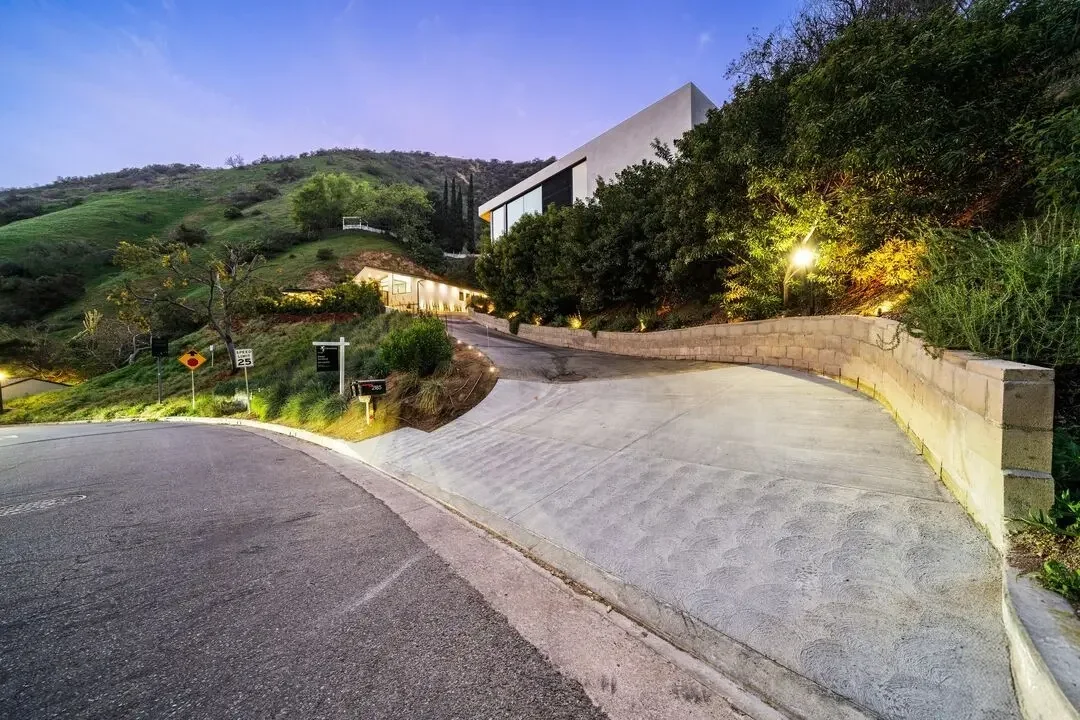 A winding driveway leading up a hillside with modern white house at the top, surrounded by greenery and trees, illuminated by outdoor lighting during twilight.