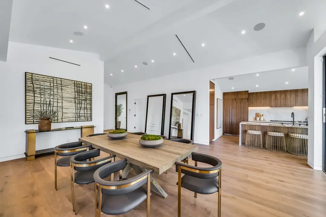 Modern dining area with wooden table, six black and gold chairs, and minimalistic artwork on the wall, open kitchen with wood cabinets and bar seating.