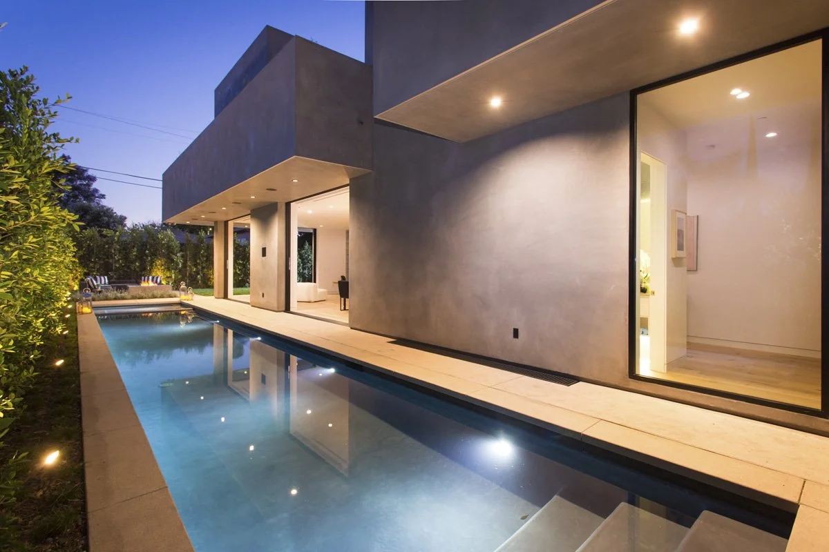 Linear lap pool running alongside minimalist plaster exterior with full-height glazing, integrated deck drains, and recessed soffit lighting.