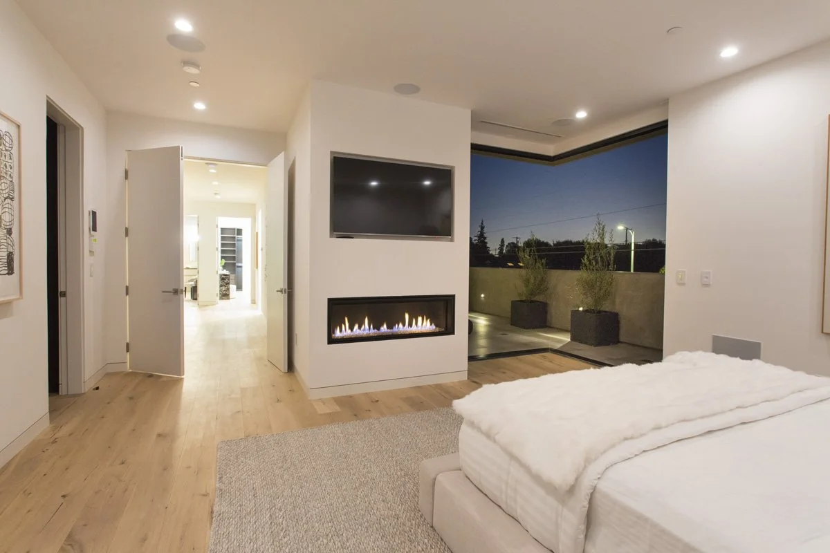 Primary bedroom with linear gas fireplace, wall-mounted television, wide-plank oak flooring, and sliding glass door opening to private terrace at dusk.