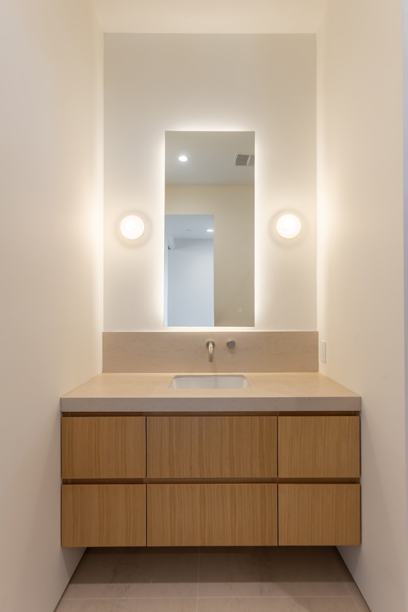 Minimalist powder room vanity with floating light oak cabinetry, integrated stone countertop and sink, wall-mounted faucet, and backlit mirror in a modern Mar Vista custom home by Heart Construction.