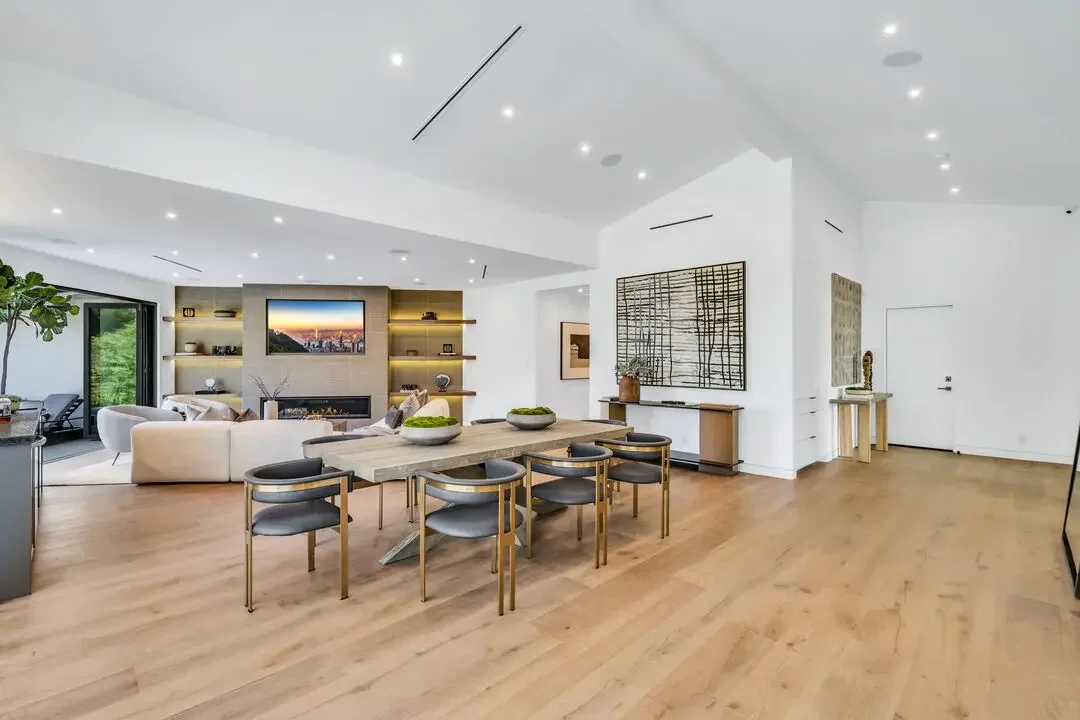 Modern living room with white walls, hardwood floors, a dining table with six chairs, a white sofa, a TV above a fireplace, and decorative shelving and artwork.