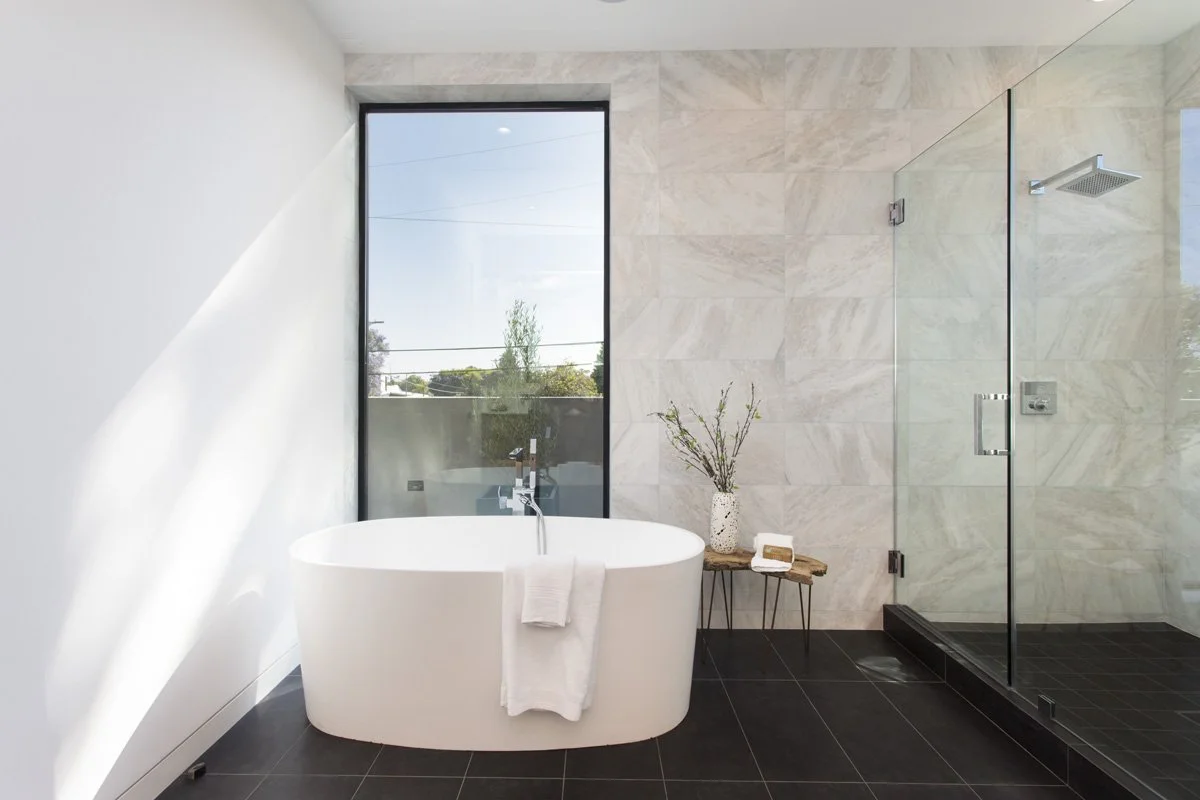 Primary bathroom with freestanding soaking tub, full-height stone tile accent wall, frameless glass shower, and matte black porcelain flooring.