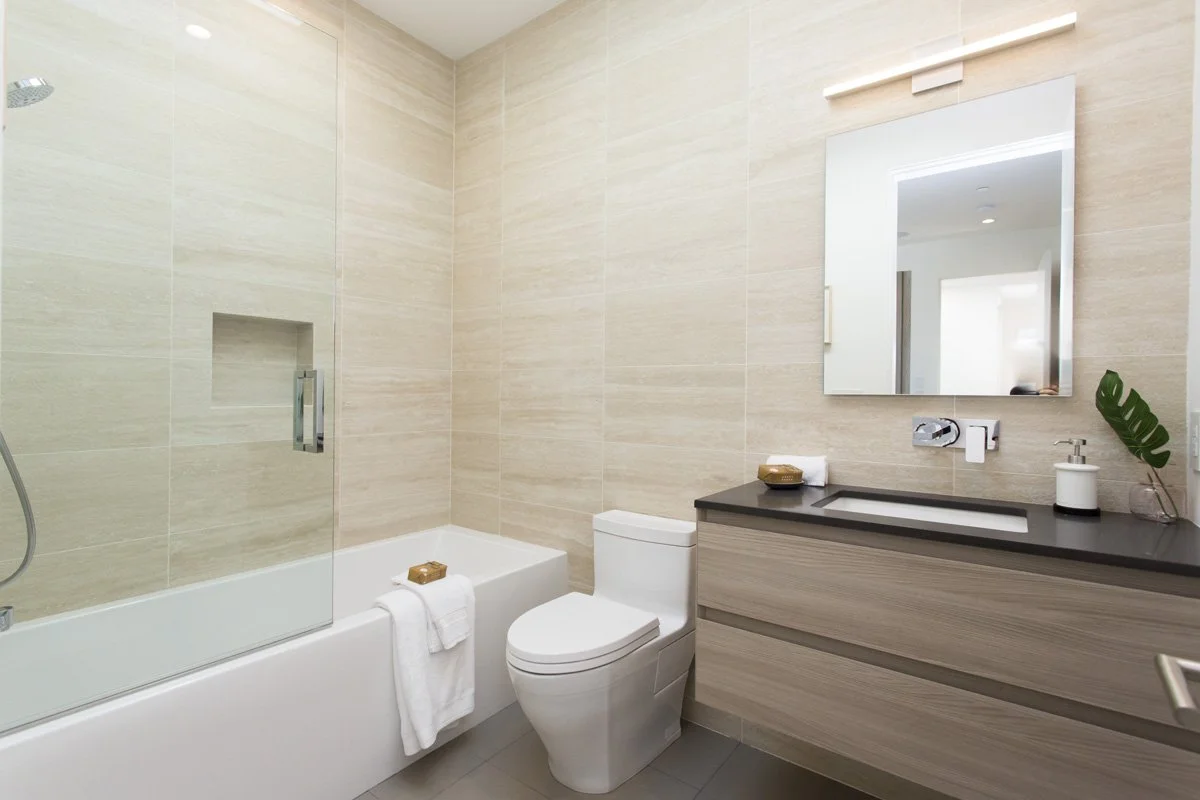 Modern bathroom with full-height beige stone tile walls, frameless glass tub enclosure, floating wood vanity with integrated sink, and wall-mounted chrome faucet.