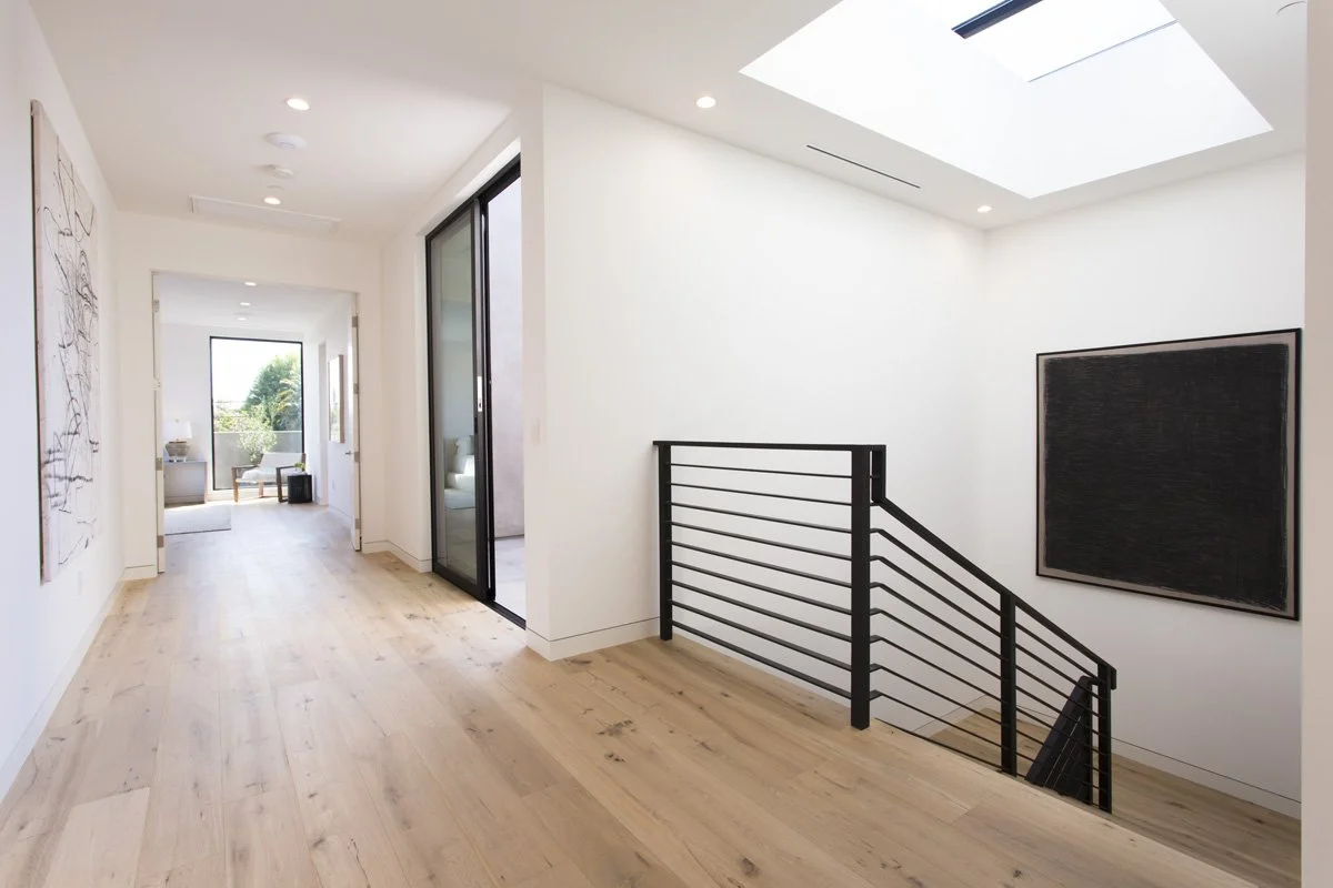 Second-floor gallery hallway with wide plank oak flooring, overhead skylight, minimalist black guardrail, and curated contemporary artwork.