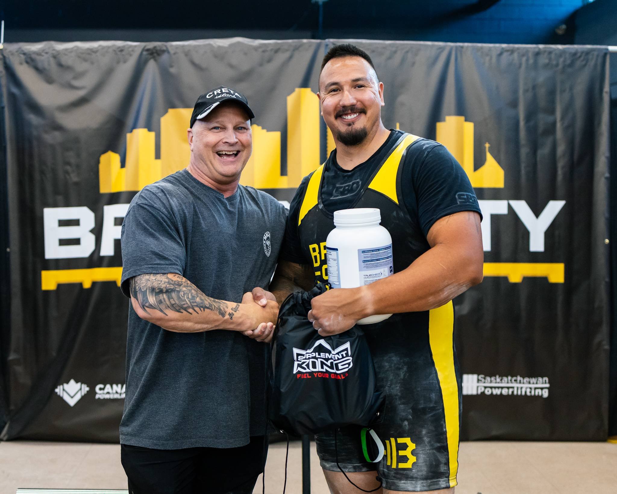 Two men smiling and shaking hands at a powerlifting event, one wearing a black and yellow singlet and holding a large container of supplement, with a banner for Saskatchewan Powerlifting in the background.