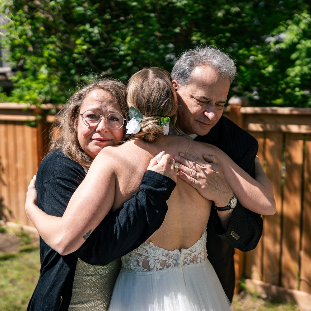 Three people hugging outdoors, including a bride in a white wedding dress, a woman with glasses, and a man in a black suit, with a wooden fence and green trees in the background.