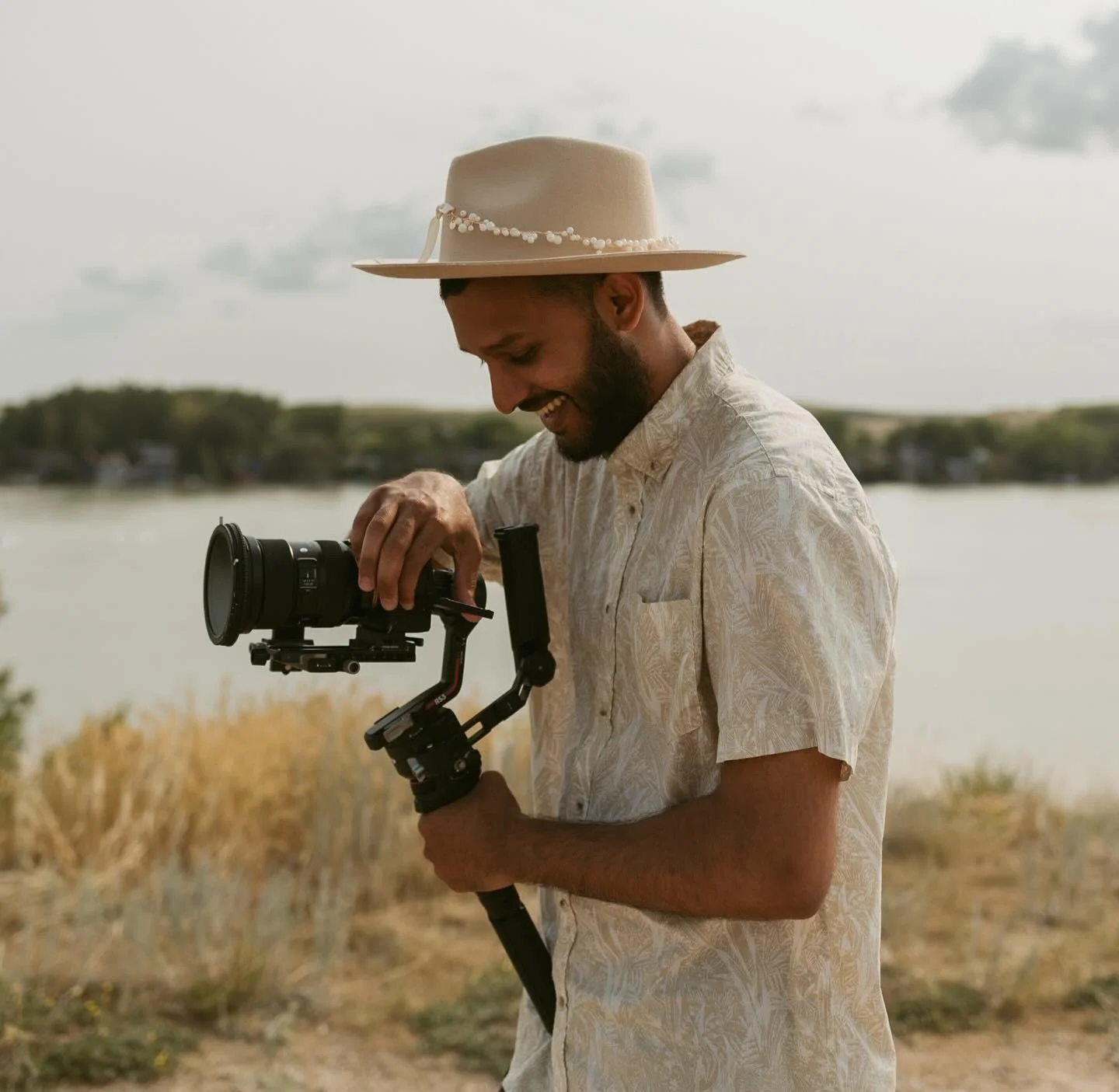 A man outdoors near a body of water, holding a camera stabilizer with a camera attached, wearing a patterned shirt and wide-brimmed hat, smiling.