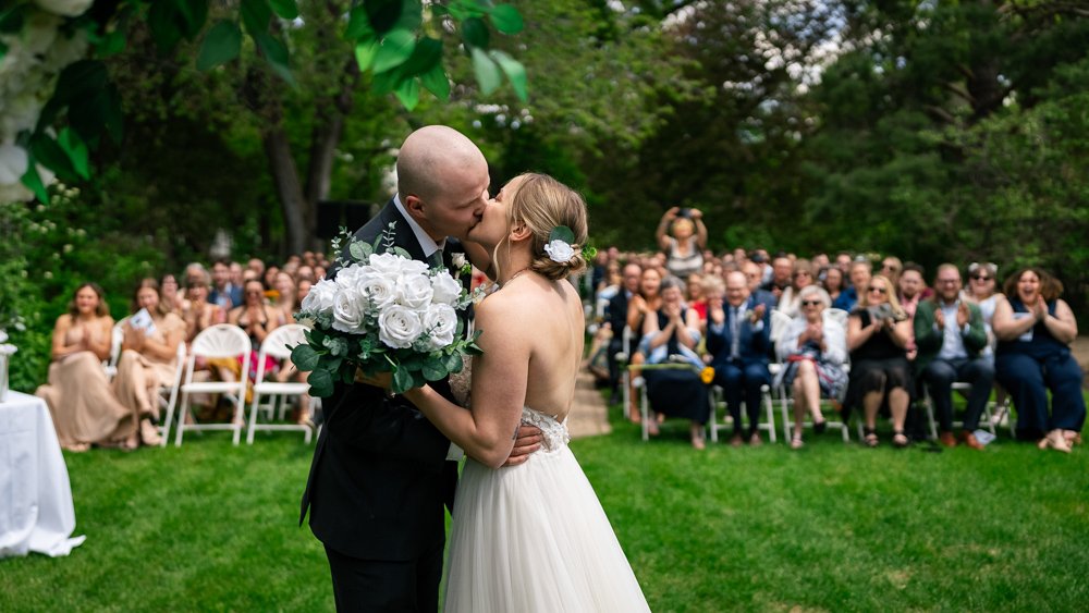 A newly married couple sharing a kiss at an outdoor wedding ceremony, with guests clapping and celebrating in the background.