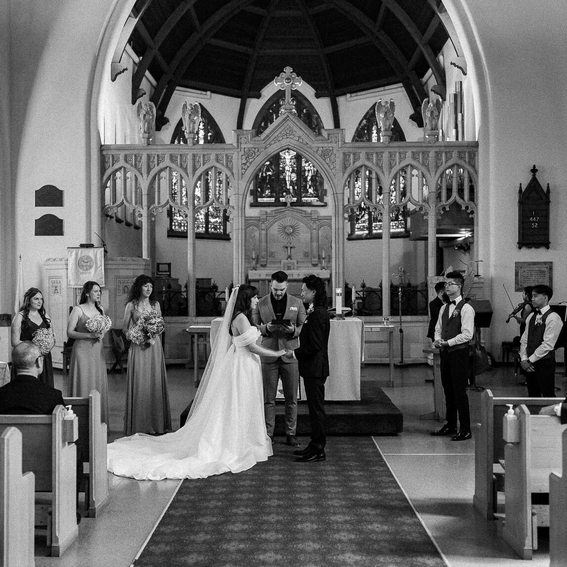 Black and white photo of a wedding ceremony inside a church with a bride and groom holding hands at the altar, surrounded by bridesmaids and groomsmen, with the officiant reading. The church has stained glass windows and wooden architectural details.