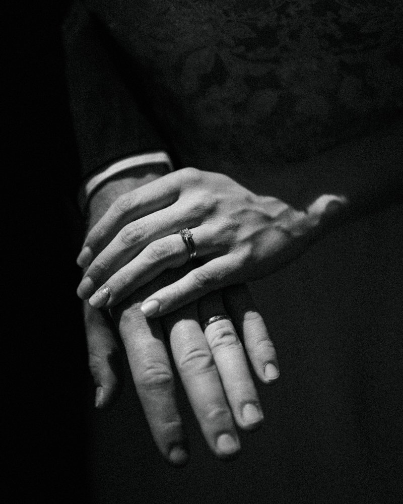 Two hands, one resting on top of the other, with rings on the ring fingers, in black and white.