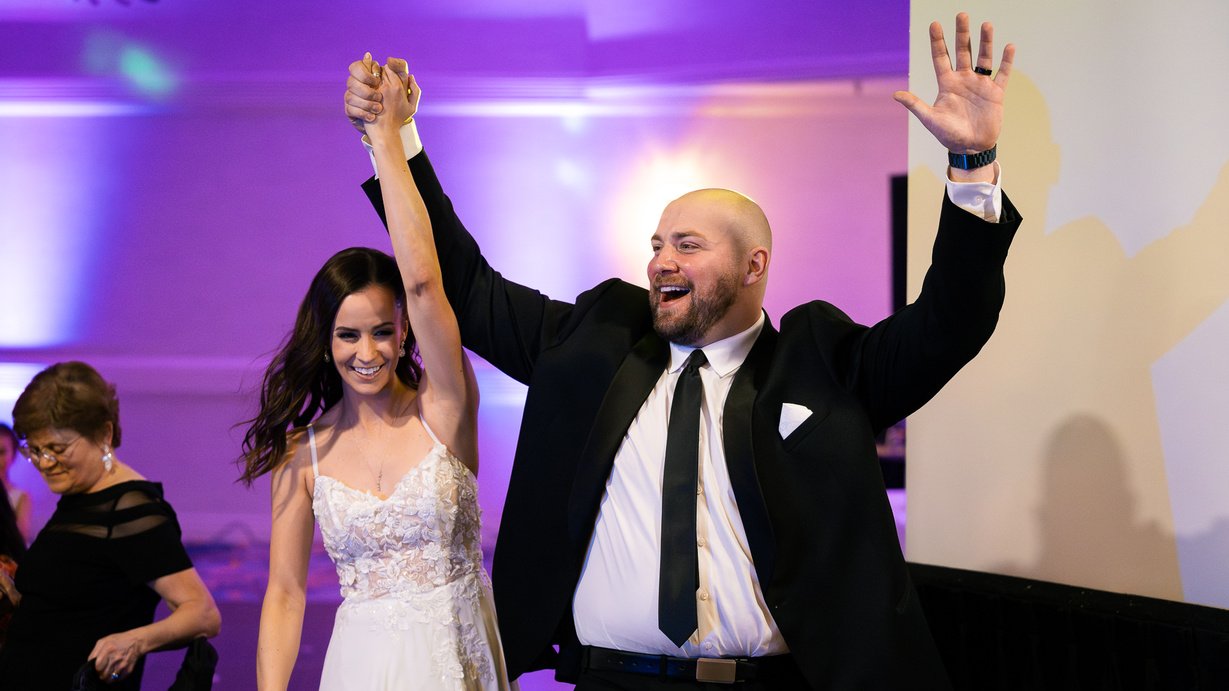 Man and woman celebrating, holding hands in the air, dressed in formal attire at a celebration or wedding reception.