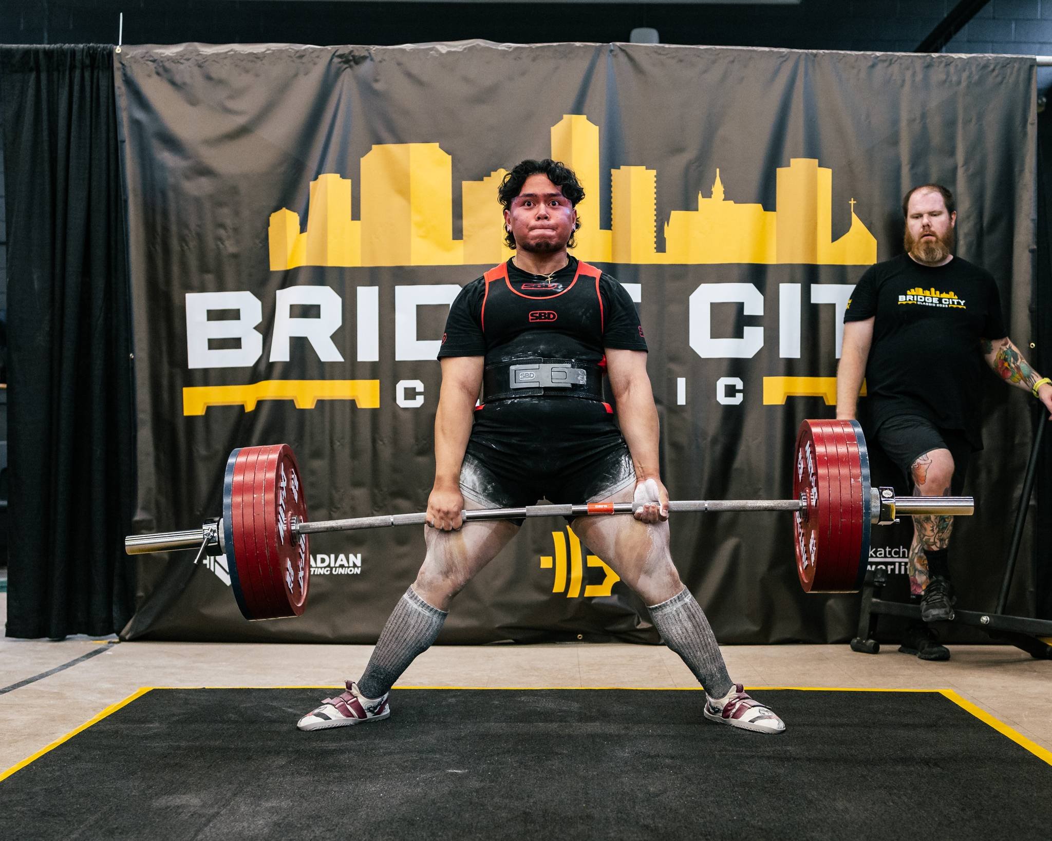 A man performing a deadlift with a loaded barbell at a powerlifting competition. He is wearing a black weightlifting suit, socks, and lifting shoes, with chalk on his hands. A judge or coach stands behind him observing.