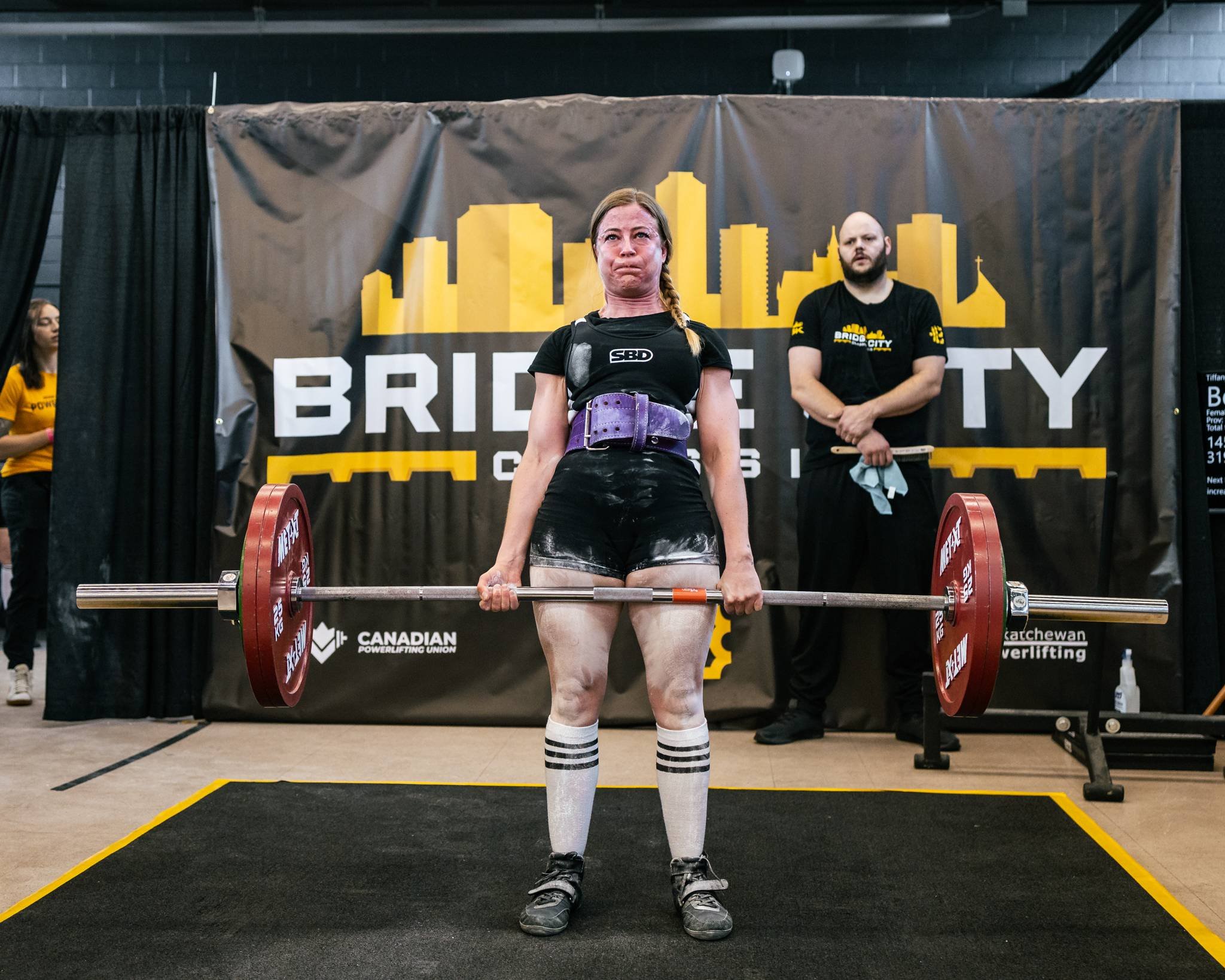 A woman performing a deadlift during a powerlifting competition, wearing a black shirt, shorts, knee-high socks, weightlifting belt, and lifting a barbell with red weights. Behind her is a black banner with yellow and white text and silhouettes of bu