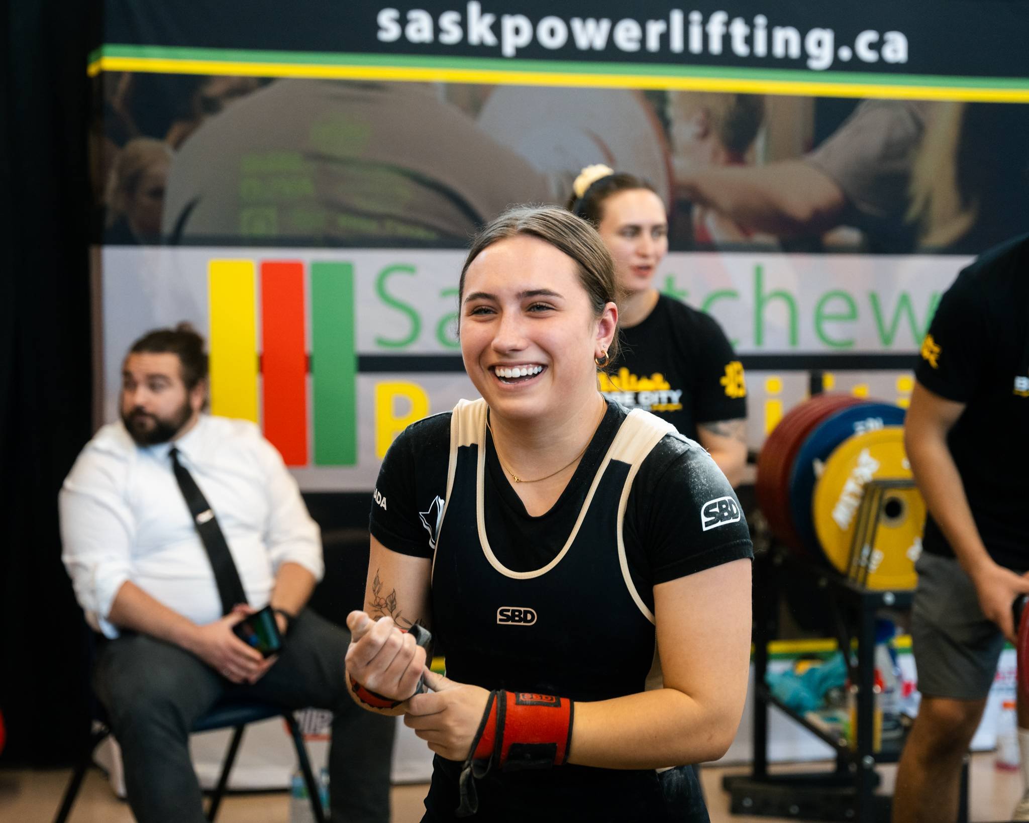 A young woman with brown hair smiling and holding a clenched fist at a powerlifting event. She is wearing a black powerlifting singlet and wrist wraps. In the background, a man in a white shirt and tie and two women standing behind her are visible, w