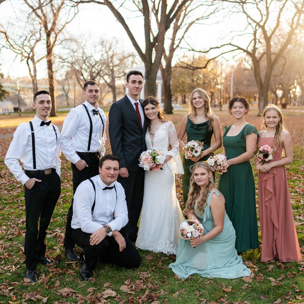 Group of people dressed for a wedding, standing outdoors in fall with trees and leaf-covered ground, during sunset.