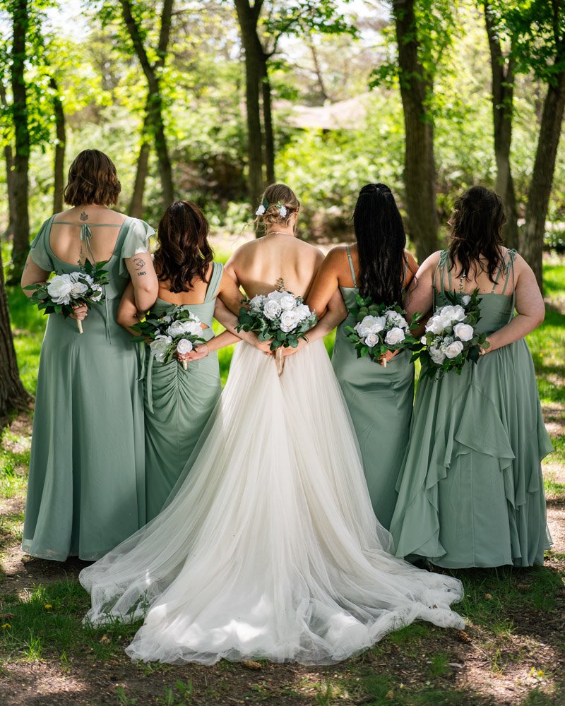 Bride and four bridesmaids standing outdoors in a wooded area, holding bouquets of white flowers, with the bride wearing a white gown and the bridesmaids in matching green dresses.