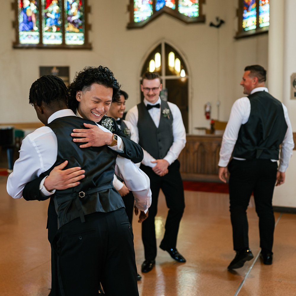 A group of five men dressed in formal wedding attire, with some hugging and smiling inside a church with stained glass windows.