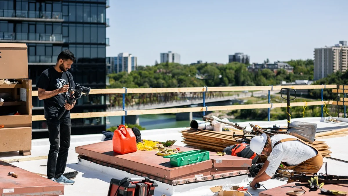 Construction workers on a rooftop with tools and materials, one worker wearing a white helmet and another guy taking photos.