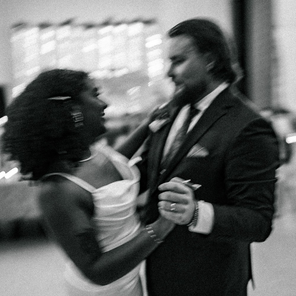 A black woman and a white man dancing in a dimly lit room, with the woman wearing a light-colored dress and the man in a suit.