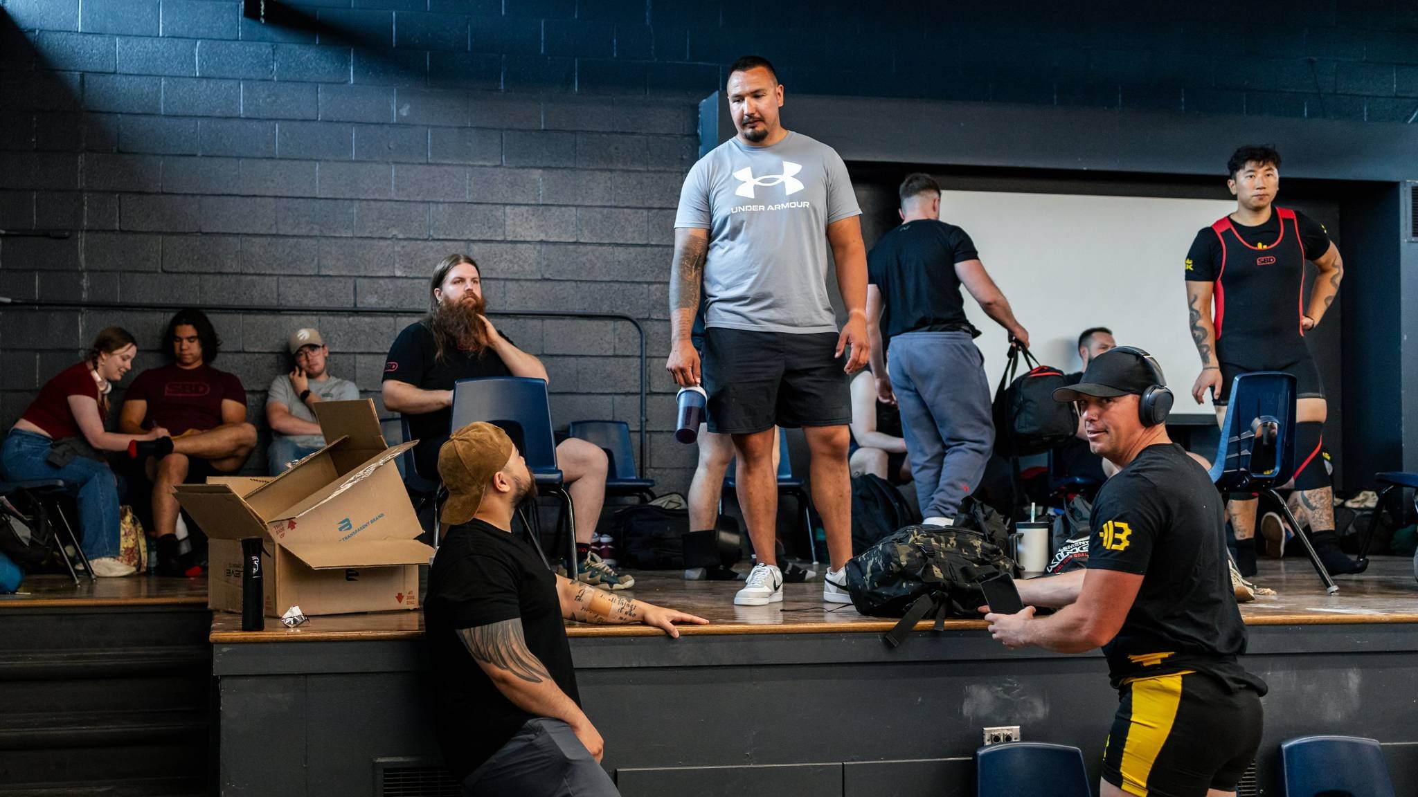 Group of people in a gym or fitness center, some sitting, some standing, with athletic equipment and backpacks, engaged in conversation or resting.