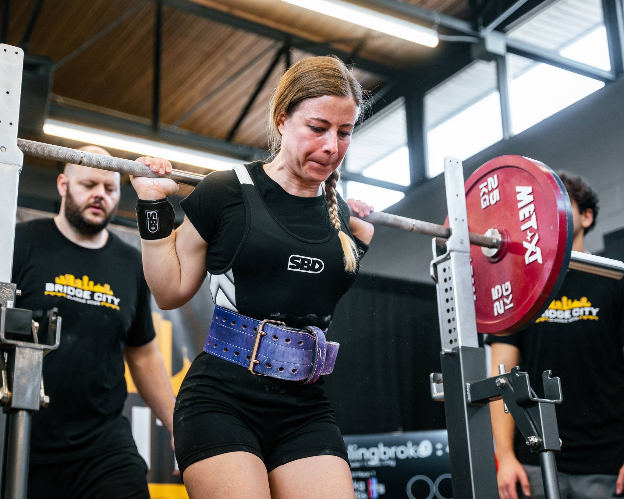 A woman performing a squat exercise with a barbell loaded with red weights in a gym, monitored by two spotters, one nearby and one in the background.