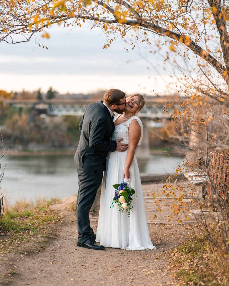 A bride and groom happily sharing a kiss on a scenic outdoor path near a river, with fall foliage and a bridge in the background.