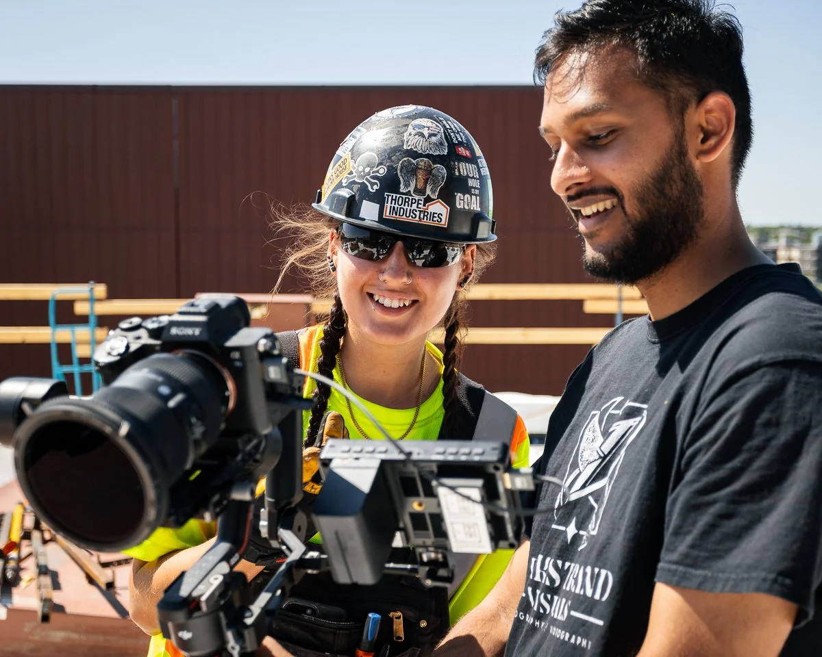 A woman wearing a construction helmet, sunglasses, and a high-visibility vest is smiling while looking at a camera mounted on a stabilizer. A man in a black t-shirt is standing beside her, also looking at the camera. They appear to be on a construction site or rooftop under a clear sky.