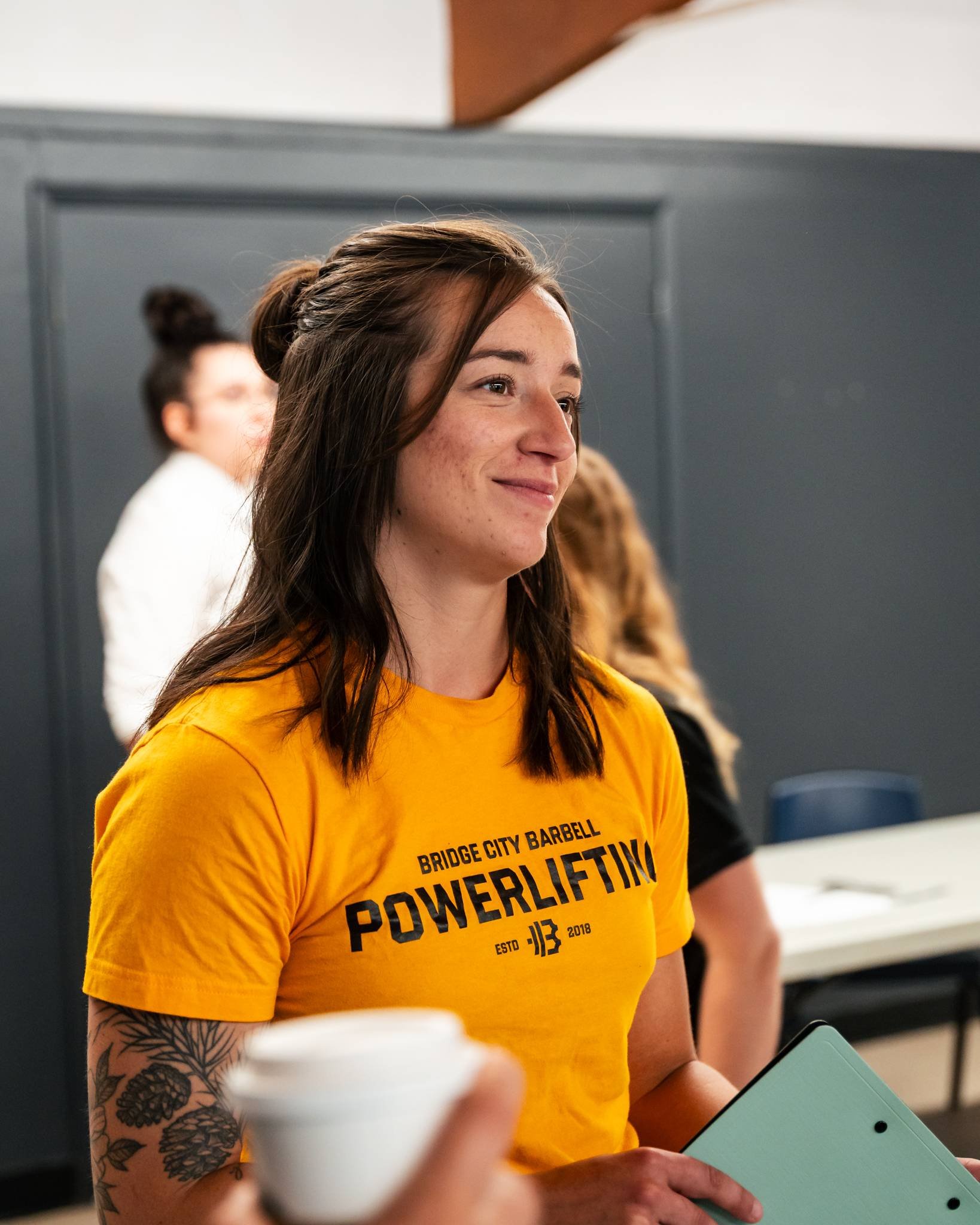 A young woman with brown hair, tattoos on her arm, wearing a yellow t-shirt with the words 'Bridge City Barbell Powerlifting' and holding a green folder, standing in a gym or fitness center.