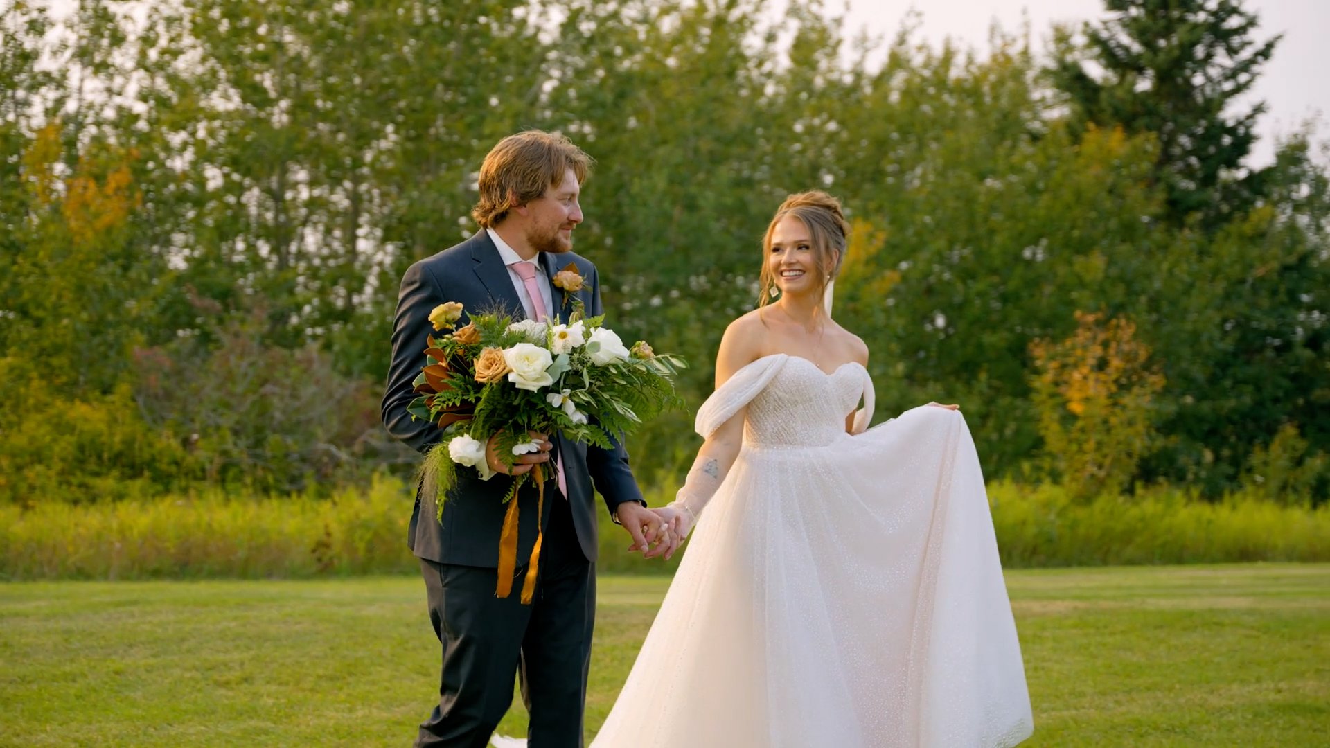 A bride and groom holding hands outdoors on their wedding day, smiling at each other, with trees in the background.
