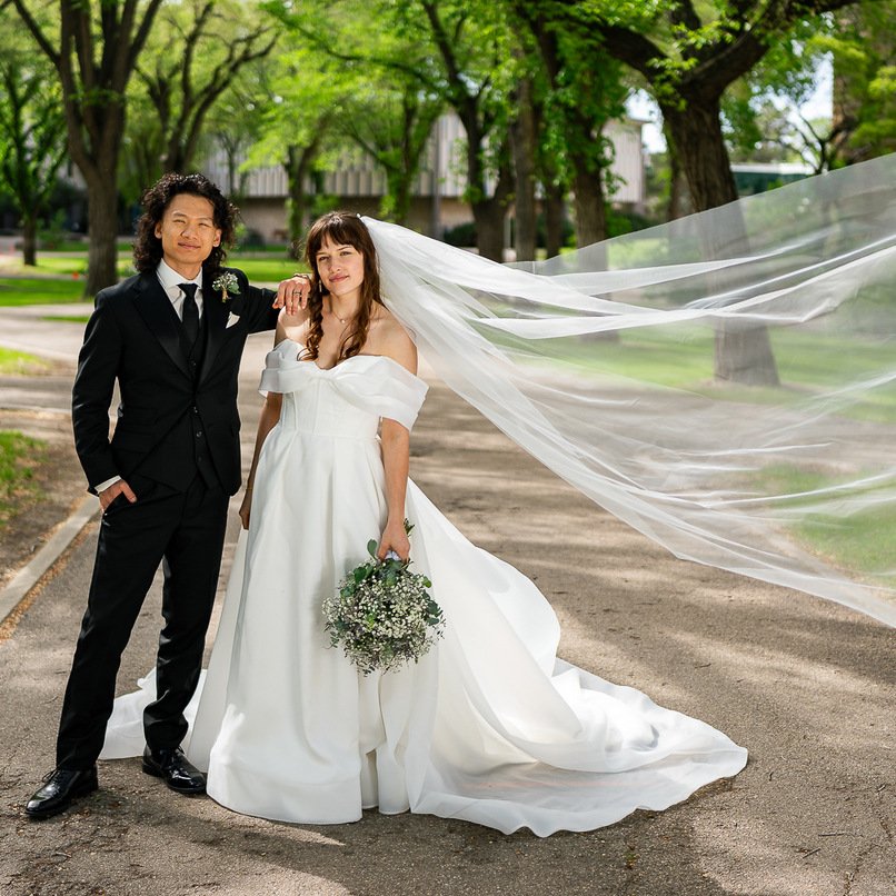 A bride and groom standing together outdoors on a sunny day, with the bride wearing a white wedding dress and veil, holding a bouquet, and the groom in a black tuxedo with a boutonnière, surrounded by green trees.
