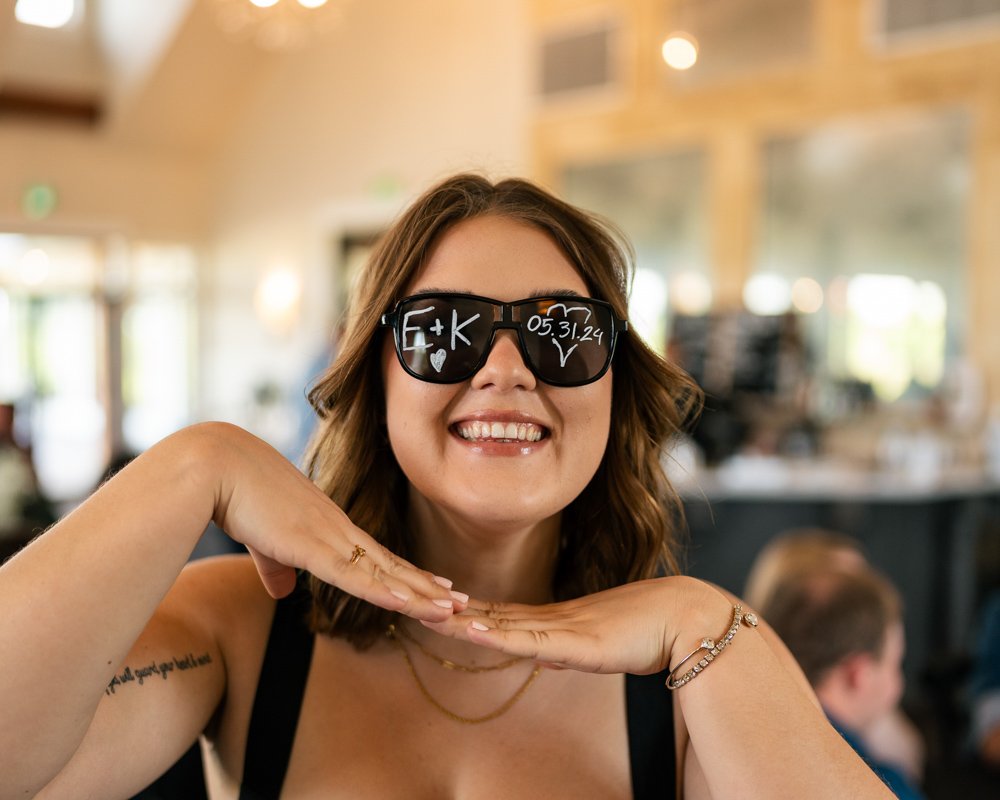 A woman wearing sunglasses with handwritten messages, smiling with her hands under her chin, in a brightly lit indoor setting.