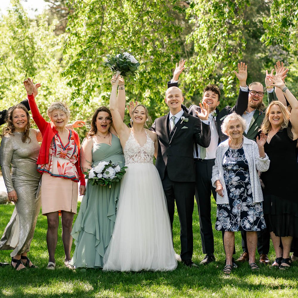 A group of people celebrating outdoors at a wedding, including a bride and groom in center, surrounded by family and friends, some raising their hands and smiling.