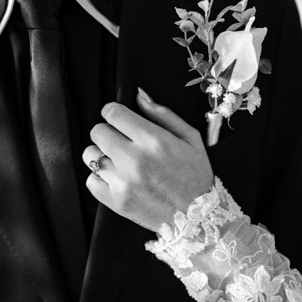 Close-up of a woman's hand with a ring, resting on a man's suit, with a boutonniere pinned to his lapel, in black and white.