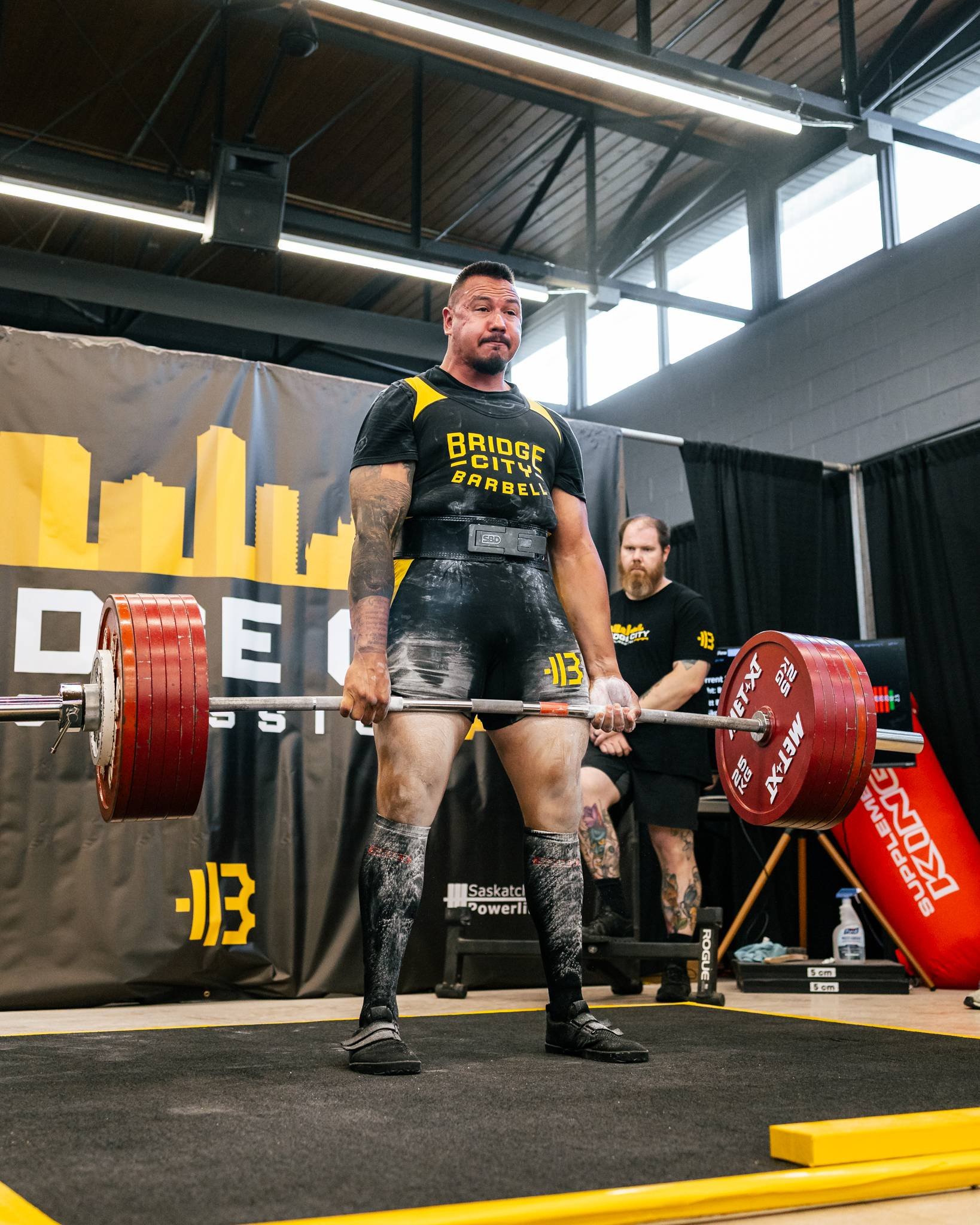 A male powerlifter in black and yellow gear performing a deadlift at a competition, with an official or coach observing in the background.