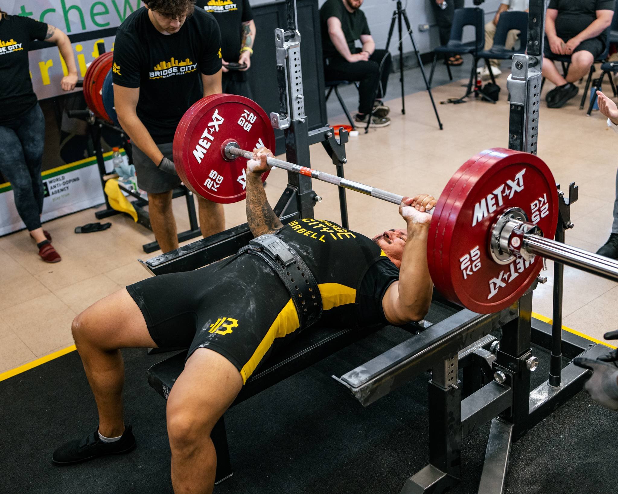 A man in black and yellow sportswear is lying on a bench, lifting a loaded barbell in a powerlifting competition. Several red weight plates are on the barbell, and his dressed in the belt and shorts with the event logo. Other people, some wearing bla