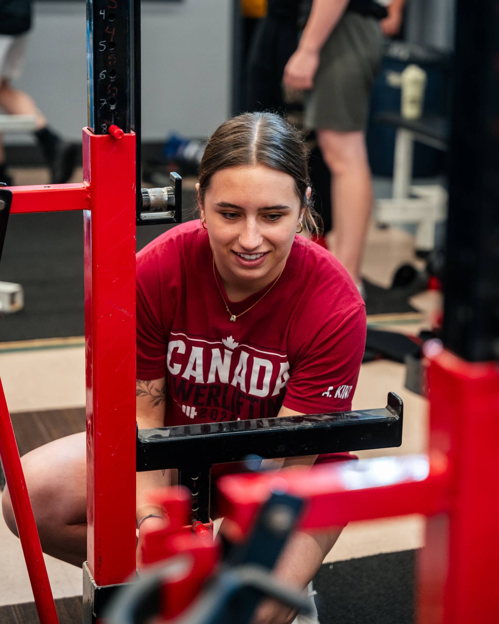 A young woman with brown hair and hoop earrings, wearing a red Canada Powerlifting T-shirt, is preparing to lift weights at a squat rack in a gym.