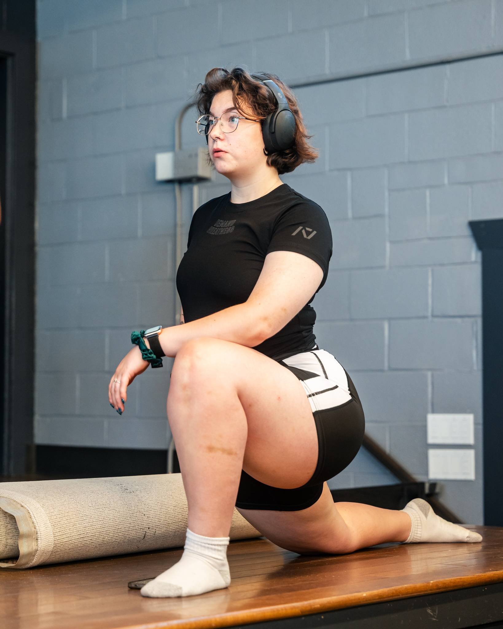 A woman with short curly brown hair, glasses, wearing a black t-shirt, black and white shorts, and white socks, kneeling on a wooden table with one knee, in a room with gray brick walls and an electrical outlet, next to a rolled-up yoga mat.